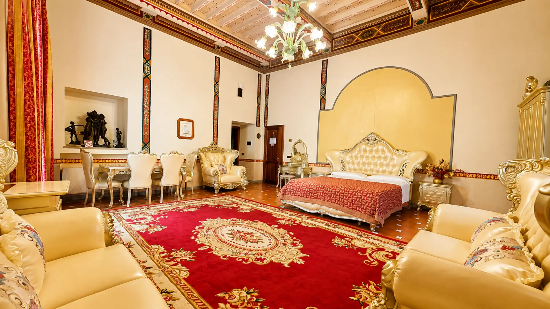 Una Junior Suite vista dall'ingresso: soffitto a travi dipinte, lampadario di vetro, tavolo da pranzo e letto matrimoniale in fondo